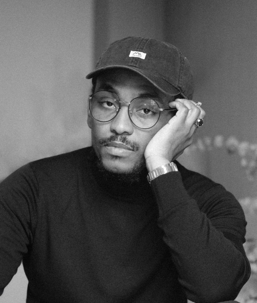 Black and white portrait of a person wearing glasses, a dark long-sleeve shirt, and a cap with a small logo. The individual is resting their head on one hand, looking thoughtful. They also have a wristwatch on the left wrist.